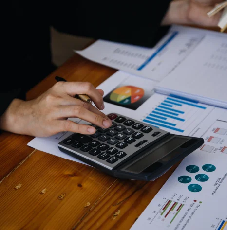 Pessoa digitando valores em uma calculadora em uma mesa de madeira com papéis com vários gráficos sobre a mesa